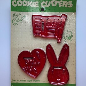 May include: A set of three red plastic cookie cutters in the shapes of a flag, a heart, and a bunny. The packaging reads "COOKIE CUTTERS" and "3 more unusual Cookie Cutters to add to your collection."