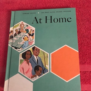 May include: A teal blue book with the title "At Home" in white text. The book is part of "The Basic Social Studies Program" and is a "Diamond Edition". The cover features illustrations of a family and a party.