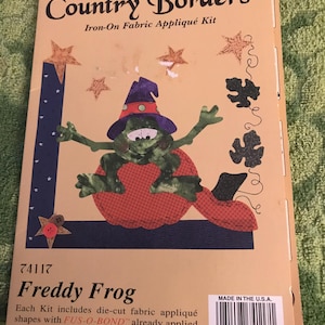May include: A craft kit for making a Freddy Frog appliqué. The kit includes die-cut fabric shapes with FUS-O-BOND™ already applied. The image shows a green frog wearing a purple and white witch hat sitting on a red and white pumpkin. The kit is part of the Harvest Country Borders collection.