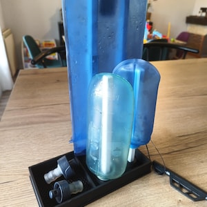 Peut inclure: Un égouttoir rectangulaire noir supportant trois poches à eau bleues de tailles différentes, avec deux bouchons en plastique gris posés dans le compartiment avant de la base, le tout présentant des gouttelettes d'eau visibles en surface.