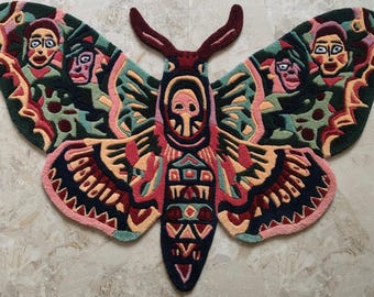 May include: A tufted rug shaped like a large moth with intricate patterns in dark green, deep red, peach, and light blue. The wings feature stylized human faces, while the central body displays geometric shapes and a skull-like motif.