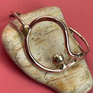 May include: A rose gold-colored bracelet with a unique, open design. The bracelet features a circular shape with a textured sphere accent. The jewelry is displayed on a light-colored stone against a pink background.