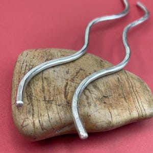 May include: Two silver-colored, wavy hair pins rest on a light brown stone against a red background. The pins have a smooth, polished finish and a gentle, curved shape, adding a touch of elegance. These hair accessories are designed to secure and style hair.