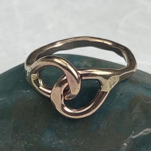 May include: A rose gold ring with a knot design. The ring features two interlocking loops, creating a continuous, intertwined pattern. The metal has a slightly hammered texture, adding visual interest to the jewelry.