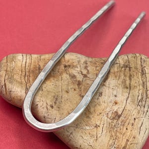 May include: A silver-colored hair pin with a hammered texture. The U-shaped pin is displayed on a light brown, textured stone against a red background. This hair accessory is a stylish addition to any hairstyle.