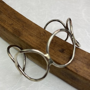 May include: A silver-colored cuff bracelet with an open, abstract design. The bracelet features multiple circular and curved elements, creating a unique, artistic look. The jewelry is displayed on a wooden surface.