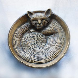 Może przedstawiać: Brązowa, okrągła miska z szczegółowym reliefem śpiącego kota zwiniętego w środku. Sierść kota i wnętrze miski mają teksturowane linie, tworzące wirujący wzór. Miska ma podniesioną krawędź.