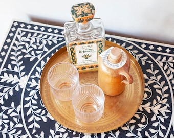 Op de afbeelding: Een gouden dienblad met twee heldere glazen en twee flessen. Eén fles is van helder glas met een groen en geel etiket waarop "Tequila Artesano" staat. De andere fles is van bruin keramiek met een wit etiket waarop "El Sabor de la Vida" staat.