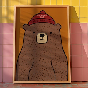 Puede incluir: Ilustración enmarcada de un oso pardo con gorro rojo con el texto "BEAR MINIMUM CLUB". El oso tiene un diseño sencillo, tipo caricatura, sobre un fondo amarillo. La obra se exhibe en una habitación con azulejos coloridos y una lavadora.