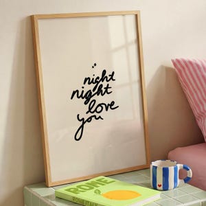 以下が含まれることがあります： 黒の手書き文字でnight night love youと書かれ、その上に小さな星が2つ描かれた縦型の額入りウォールアートプリント。プリントは明るい木製のフレームに入っており、レモンのグラフィックが描かれた緑色の本と、青いストライプのマグカップの隣に置かれています。