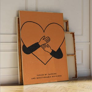 Könnte beinhalten: Gerahmter Kunstdruck mit einem Herz, das von Armen umarmt wird, vor einem strukturierten, orangefarbenen Hintergrund. Das Kunstwerk enthält den Text "FUELED BY CAFFEINE AND QUESTIONABLE DECISIONS" in minimalistischem Stil. Der Druck wird mit anderen gerahmten Leinwänden ausgestellt.