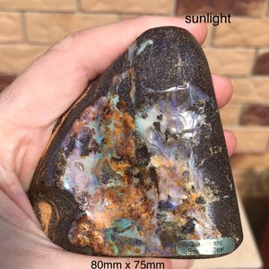 Puede incluir: Ópalo de roca de Queensland pulido, de 8 cm x 7,5 cm. La piedra muestra una matriz marrón oscuro con colores internos vibrantes de azul, morado, naranja y verde. La palabra "sunlight" es visible en la parte superior.
