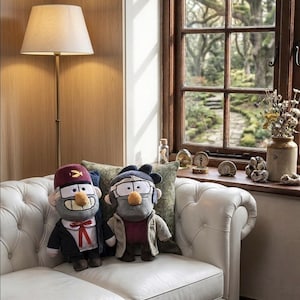 May include: Two plush character dolls from the animated series, Gravity Falls, sit on a white leather couch. One doll wears a maroon hat and a black suit with a red bow tie. The other doll wears a gray jacket and a maroon shirt. A floor lamp and window are in the background.