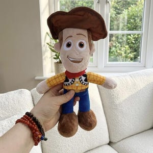 May include: A plush Woody doll from the Toy Story franchise. The doll features a brown cowboy hat, a yellow plaid shirt, blue pants, and brown boots. It has a red neckerchief and a smiling face.