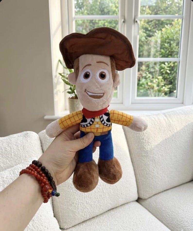 May include: A plush Woody doll from the Toy Story franchise. The doll features a brown cowboy hat, a yellow plaid shirt, blue pants, and brown boots. It has a red neckerchief and a smiling face.
