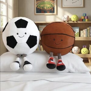 May include: A white and black soccer ball plush toy and a brown basketball plush toy with smiling faces and attached legs. The toys are on a white pillow, with other sports-themed toys in the background.