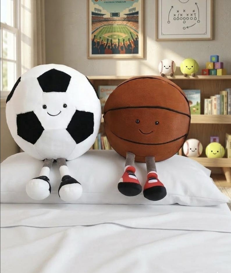May include: A white and black soccer ball plush toy and a brown basketball plush toy with smiling faces and attached legs. The toys are on a white pillow, with other sports-themed toys in the background.
