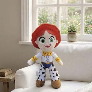 May include: A plush doll of a character with red hair, a white shirt with yellow accents, blue jeans, and black and white spotted pants. The doll has green eyes and a smiling expression. The doll is standing on a white couch in front of a window.