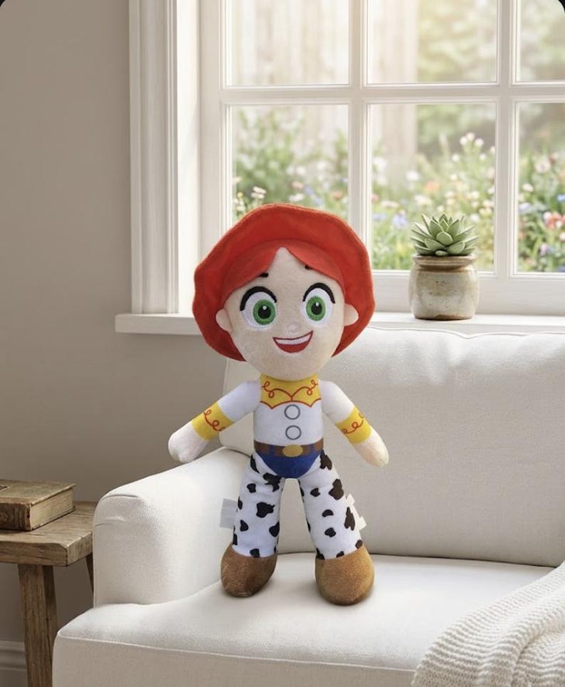 May include: A plush doll of a character with red hair, a white shirt with yellow accents, blue jeans, and black and white spotted pants. The doll has green eyes and a smiling expression. The doll is standing on a white couch in front of a window.