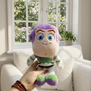 May include: A plush Buzz Lightyear doll, featuring a purple head, blue eyes, and a smiling expression. The doll wears a green and white space suit with purple and green accents. The doll is held in front of a window with a view of greenery.