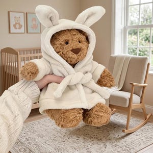 May include: A plush brown teddy bear wearing a cream-colored robe with bunny ears. The bear has a brown nose and is being held in front of a nursery setting with a crib, rocking chair, and framed artwork.