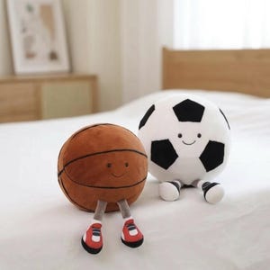 May include: Two plush sports balls with smiling faces. A brown basketball with black lines and red and white shoes sits next to a white soccer ball with black pentagons and black and white shoes. Both are on a white surface.