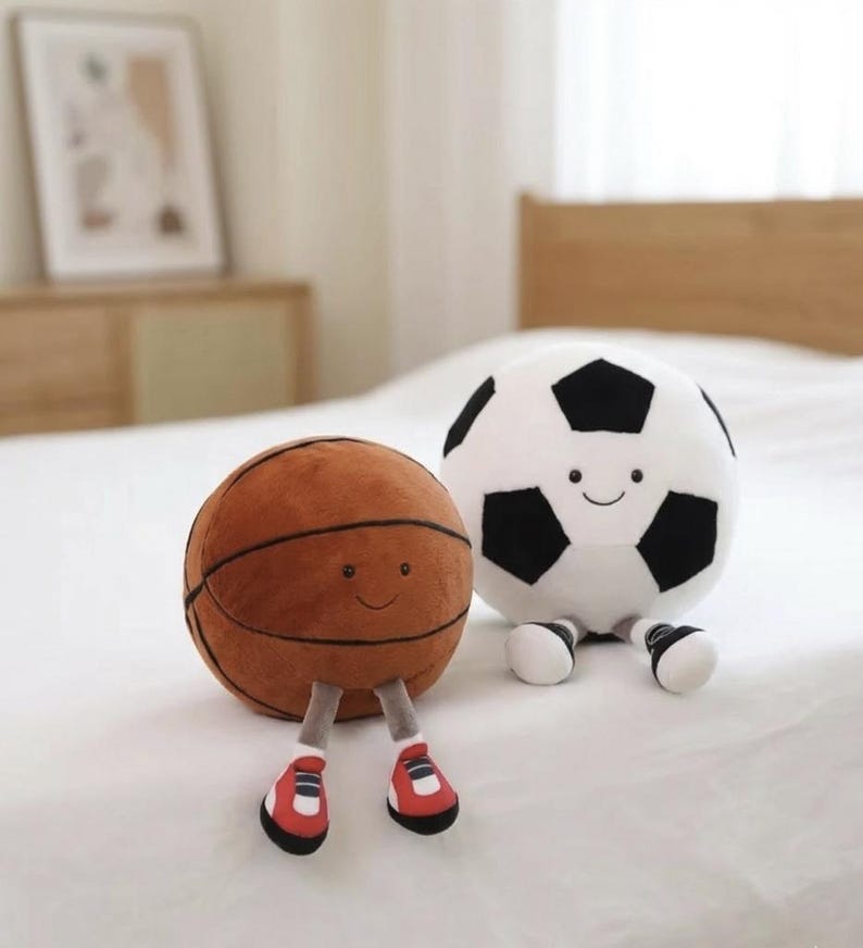 May include: Two plush sports balls with smiling faces. A brown basketball with black lines and red and white shoes sits next to a white soccer ball with black pentagons and black and white shoes. Both are on a white surface.