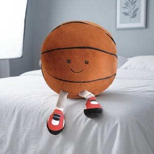 May include: A plush basketball toy with a smiling face, black stripes, and attached legs wearing red and black sneakers. The toy is brown and sits on a white bedspread.