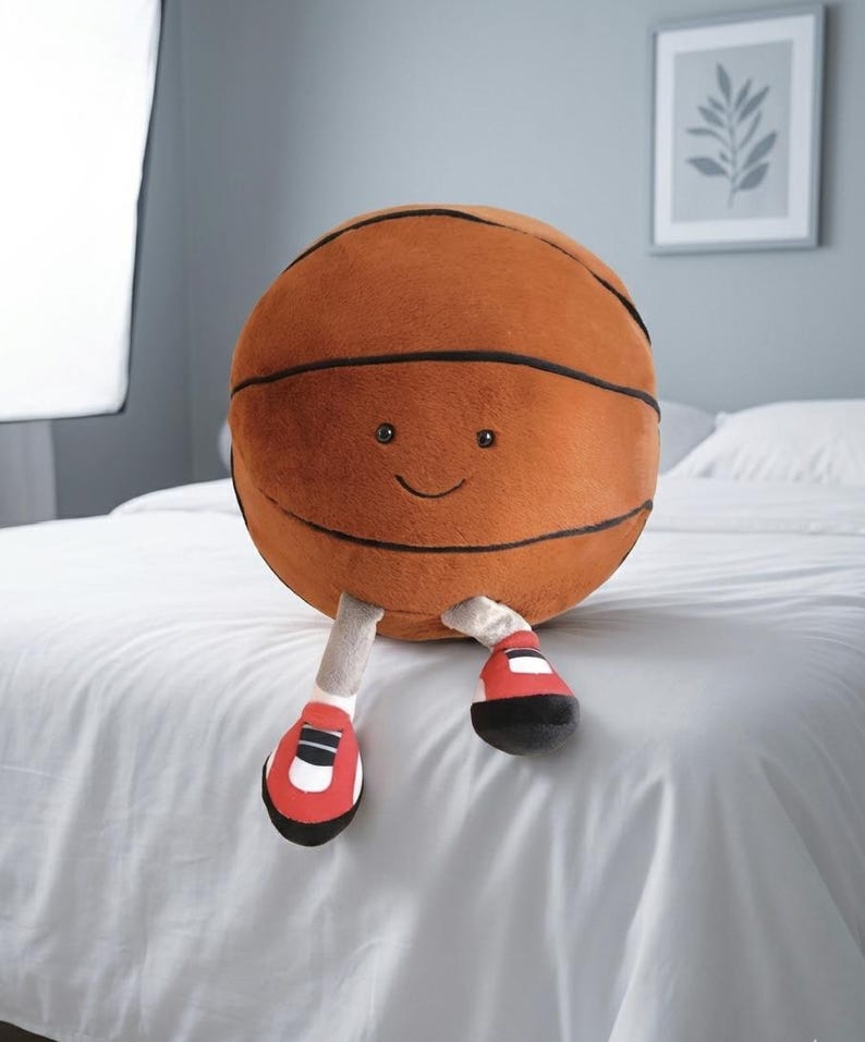 May include: A plush basketball toy with a smiling face, black stripes, and attached legs wearing red and black sneakers. The toy is brown and sits on a white bedspread.