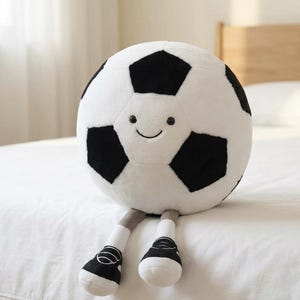 May include: A plush soccer ball with a smiling face, black and white pentagon panels, gray legs, and black and white shoes. The soft toy is designed to sit upright.