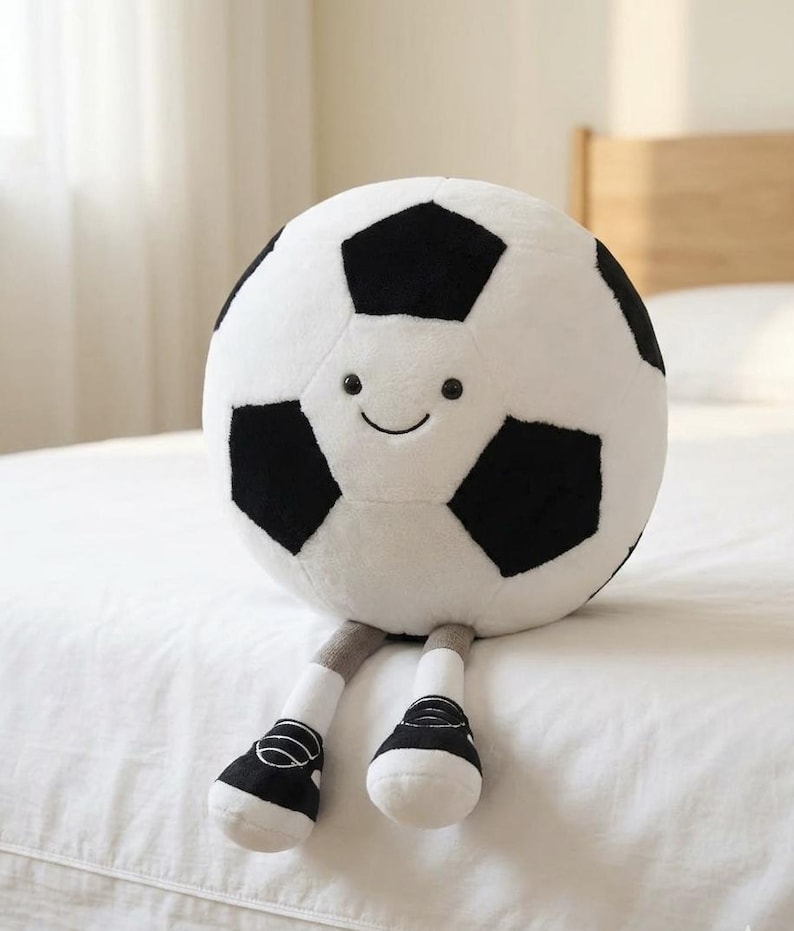 May include: A plush soccer ball with a smiling face, black and white pentagon panels, gray legs, and black and white shoes. The soft toy is designed to sit upright.