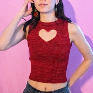 May include: A woman wearing a red knitted crop top with a heart-shaped cutout. The top has a ribbed texture and is sleeveless. She is wearing blue jeans and has her hair pulled back.