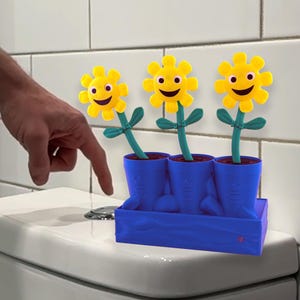 Puede incluir: Un ambientador de inodoro novedoso con tres cabezas de flores amarillas sonrientes, tallos y hojas verdes. Las flores están en macetas azules en forma de bota sobre una bandeja azul. El producto está colocado en un inodoro blanco.