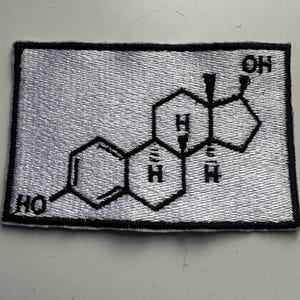 May include: A rectangular embroidered patch featuring a black and white chemical structure diagram. The design includes hexagonal shapes and chemical notations, with a white background and a black border.