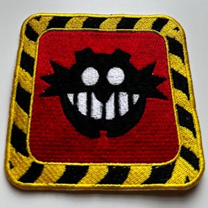 May include: Embroidered patch featuring a black and white character design on a red background, framed by a yellow and black striped border. A second, avocado-shaped patch with a smiling face is also visible.
