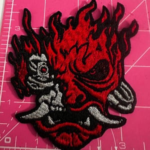 Puede incluir: Parche bordado con un rostro de demonio rojo y negro estilizado con llamas. El parche tiene detalles en blanco y negro y está colocado sobre una esterilla de corte rosa. El diseño es detallado e intrincado.