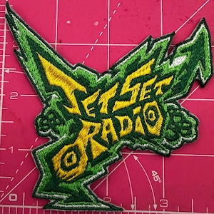 Pode incluir: Patch bordado com o texto "Jet Set Radio" em amarelo e verde. O patch tem um design irregular e estilizado com uma borda verde. O fundo é uma base de corte rosa.