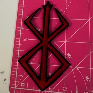 May include: A red and black embroidered patch featuring a stylized symbol. The patch has a black border and is set against a pink cutting mat with measurement markings in inches.