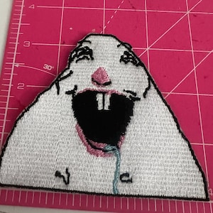 May include: Embroidered white patch featuring a cartoon rabbit with a wide open black mouth, pink nose, and drool. The patch has a black border and is placed on a pink cutting mat with grid lines.