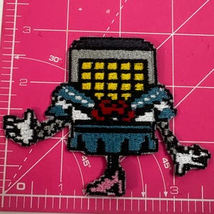 May include: A pixelated patch featuring a robot-like character with a gray head, yellow grid face, and blue and white patterned torso. The character has outstretched arms and pink feet. The patch is on a pink surface.