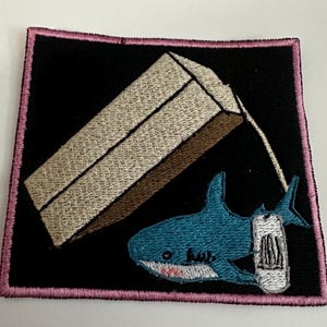 Peut inclure: Écusson brodé représentant un requin de dessin animé avec un pansement et une forme géométrique. L'écusson a un fond noir avec une bordure rose. Le requin est bleu avec un pansement blanc.