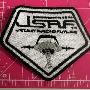 May include: Embroidered patch with the text "JSRF JET SET RADIO FUTURE" in white on a black and white pentagon shape. The patch features a gray graphic of a futuristic device. The patch is approximately 3 inches wide.