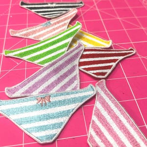 Puede incluir: Colección de parches bordados con forma de ropa interior a rayas en varios colores, incluyendo negro, rosa, verde, amarillo, rojo y azul. Algunos parches presentan un pequeño lazo. Los parches están dispuestos sobre una superficie rosa.