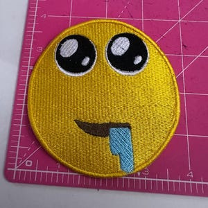 May include: Embroidered yellow patch featuring a crying emoji face with large black eyes, a brown mouth, and a blue tear. The patch is round and has a stitched border. The background is a pink cutting mat.