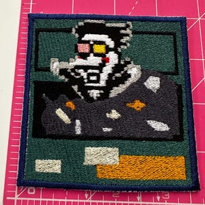 May include: Embroidered patch with a pixelated character design. The patch features a green background, a dark blue border, and a character with a white face, black hair, and yellow and pink glasses. There is a yellow and orange section at the bottom.
