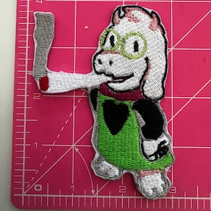 Peut inclure: Écusson brodé représentant un personnage de dessin animé avec une fourrure blanche, des lunettes vertes et une tunique verte. Le personnage tient un objet gris avec une pointe rouge. L'écusson a des accents noirs et roses sur une surface rose.