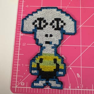 Puede incluir: Parche bordado con una figura pixelada con forma de alienígena, con una cabeza grande, dos ojos negros y un torso amarillo y negro. El parche tiene un borde azul y está sobre una esterilla de corte rosa.