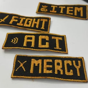 Op de afbeelding: Vier zwart en goud geborduurde patches. De patches tonen de woorden "FIGHT", "ITEM", "ACT" en "MERCY" in gouden letters. Elke patch heeft een gouden rand. De "FIGHT" patch heeft een zwaard icoon, en de "ACT" patch heeft een geluidsgolf icoon.