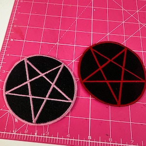 Può includere: Due toppe nere ricamate con disegni di pentagrammi. Una toppa ha un contorno rosa e un pentagramma rosa, l'altra ha un contorno rosso e un pentagramma rosso. Le toppe sono rispettivamente circolari e ovali.