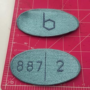 May include: Two oval-shaped, teal embroidered patches. One patch features the letter "b" inside a hexagon. The other patch has the numbers "8872" embroidered on it. The patches are on a pink cutting mat.